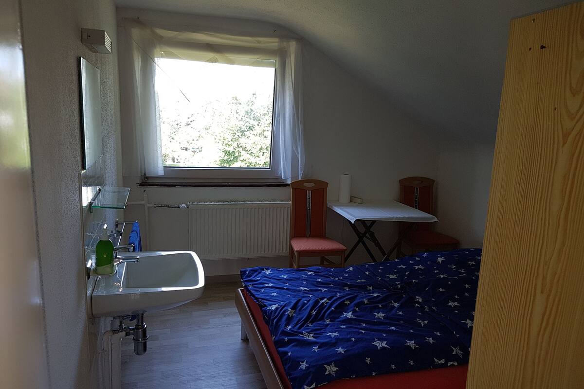 Ein kleiner, kompakter Raum mit einem Bett, einem kleinen Tisch, einem Stuhl, einem Fenster mit Vorhängen, einem Heizkörper und einem Waschbecken.