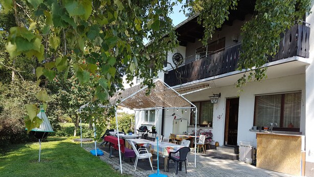 A covered outdoor dining area with tables and chairs, a barbecue grill, and a garden with a lawn and trees. - Gasthof Waldeck (Engstingen)