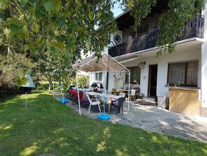 A covered outdoor dining area with tables and chairs, a barbecue grill, and a garden with a lawn and trees.