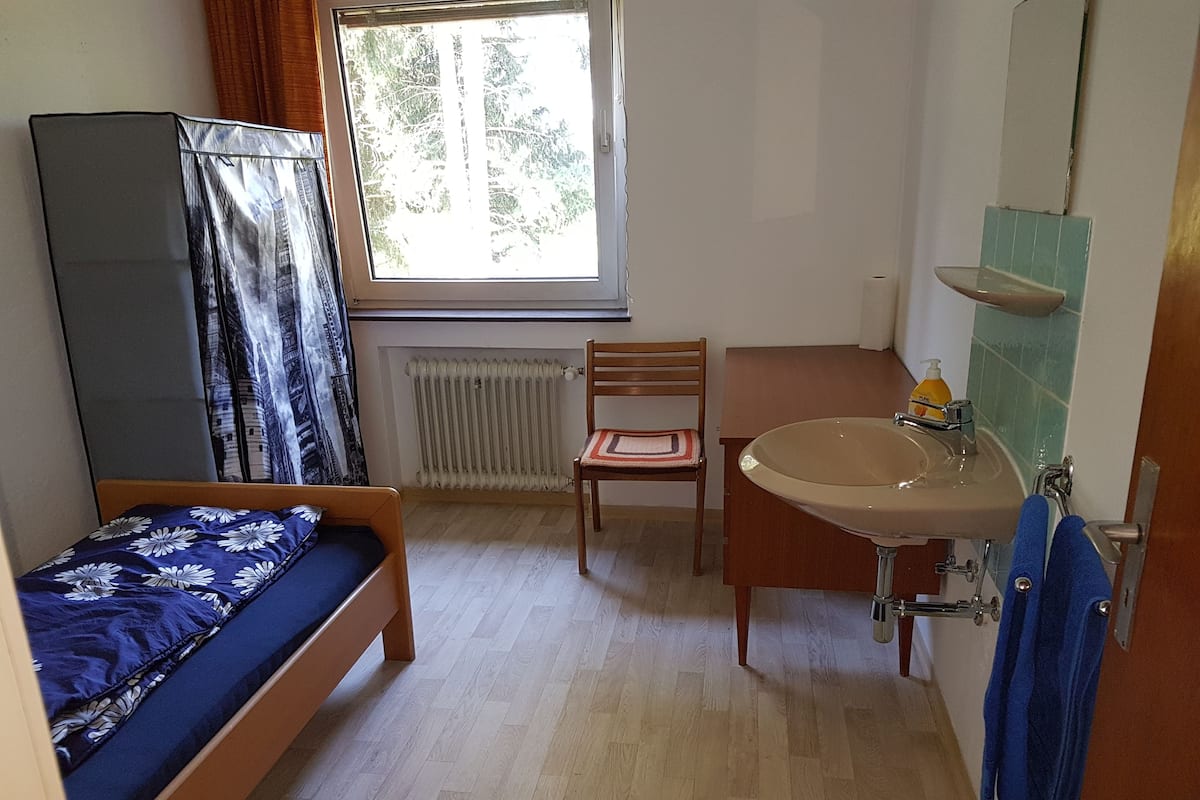 Ein kleines, gut beleuchtetes Zimmer mit einem Bett, einem Sessel, einem Waschbecken und einem Fenster.