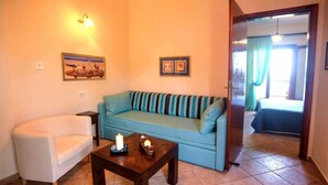 A living room with a sofa, a small table with candles, and a chair. There are framed pictures on the wall and a door leading to another room. - Anna Karra Studios & Apartments (Sithonia)