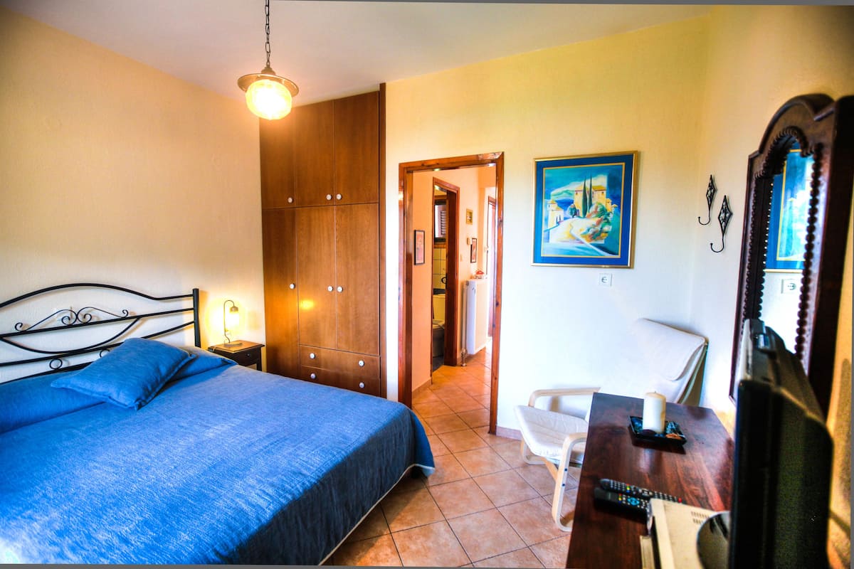 Ein Schlafzimmer mit einem Bett, einem Schreibtisch mit Stuhl, einer Schreibtischlampe, einem Gemälde und einem Spiegel.