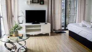 Une chambre d’hôtel moderne avec un lit, un téléviseur posé sur un meuble blanc, une table en verre agrémentée d’une plante, et une vue sur la ville grâce à de grandes fenêtres.
