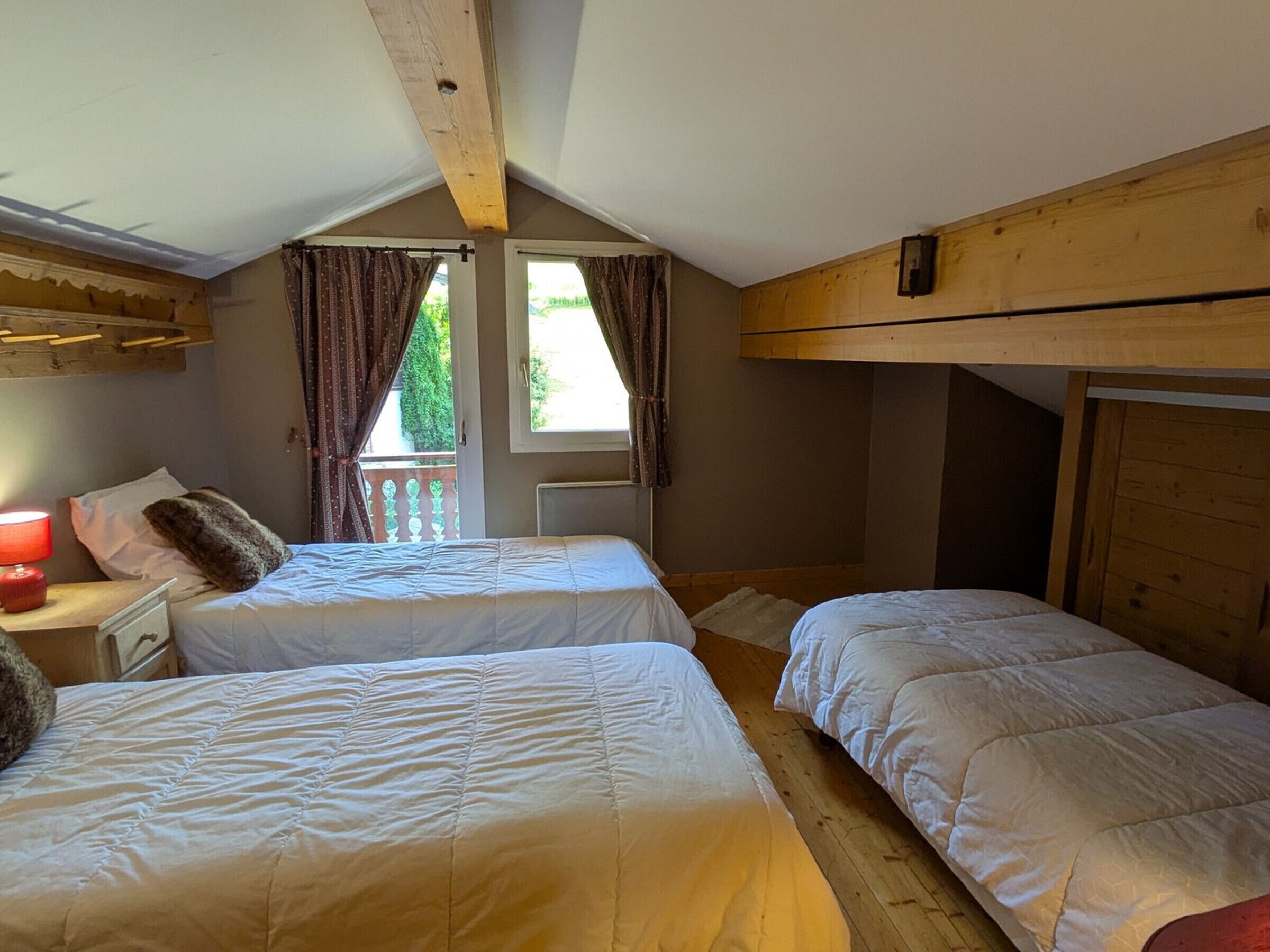 Ein Zimmer mit zwei Betten, einem Vorhangfenster, einer Holzbalkendecke und einer roten Lampe links.