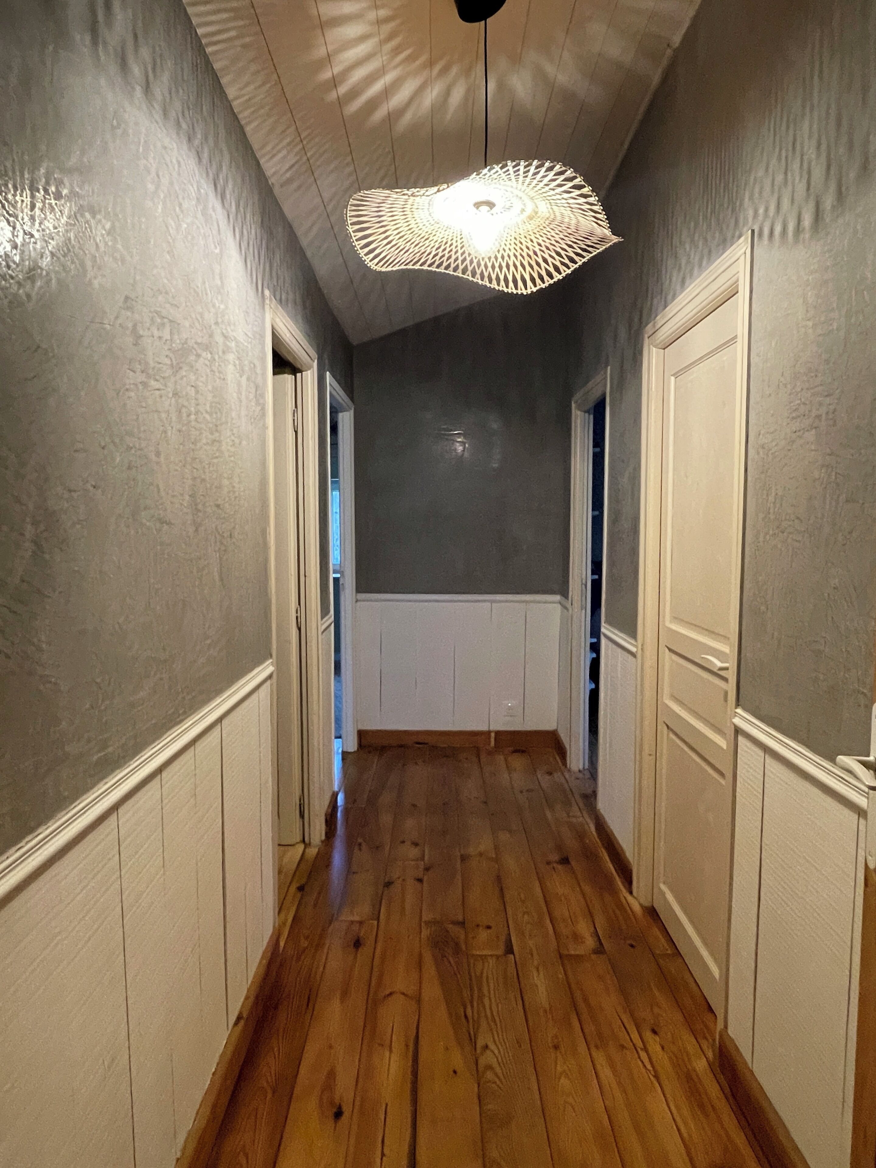 Un couloir doté d’un plancher en bois, de lambris blancs et d  ’un luminaire au plafond.