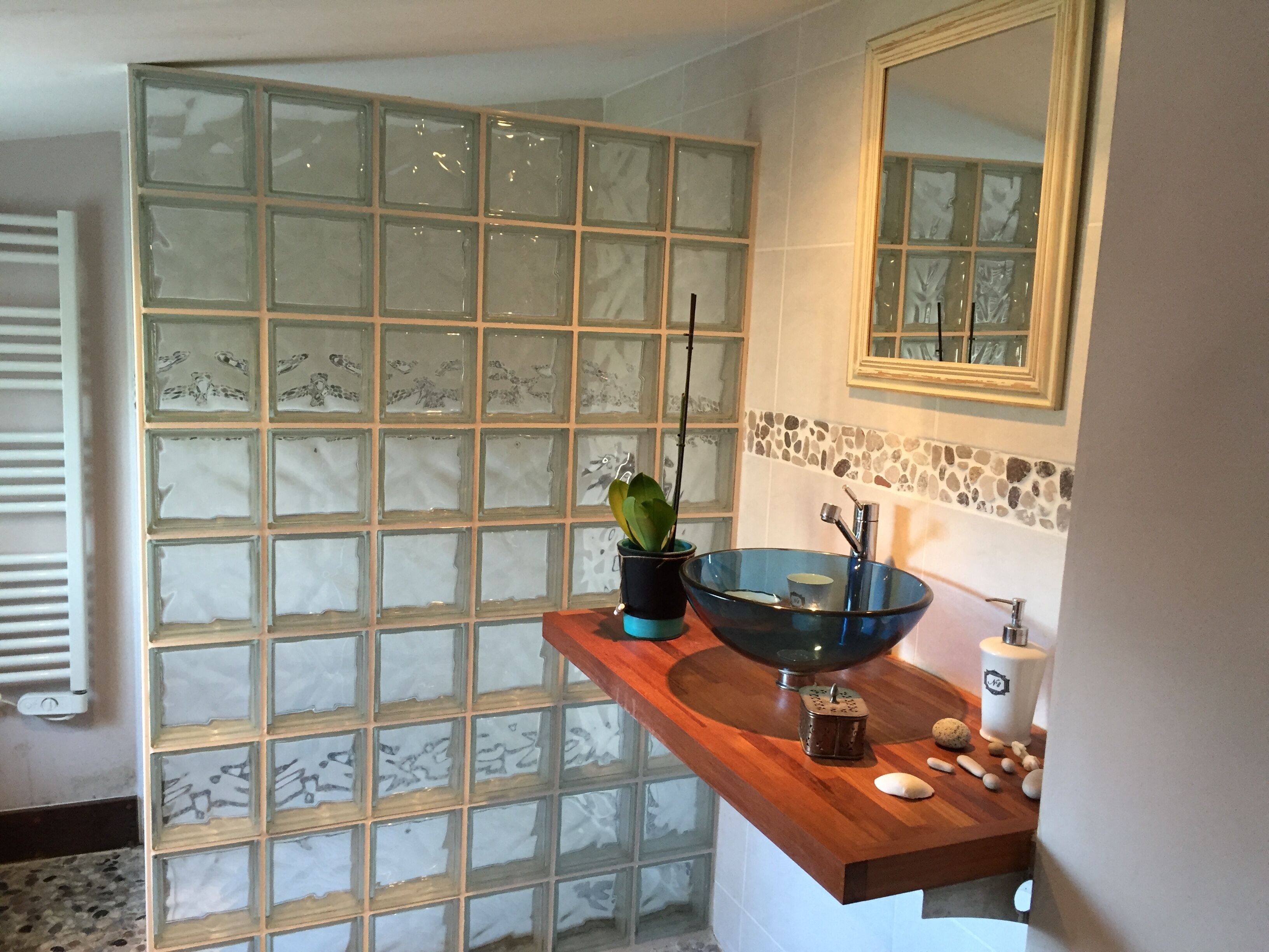 Un baño con separador de bloques de vidrio, encimera de madera, lavabo de vidrio azul, espejo y salpicadero con mosaico de piedra.