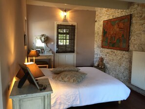 Un dormitorio con cama, escritorio con lámpara, cómoda, ventana con persianas, radiador fijado a la pared y un cuadro.