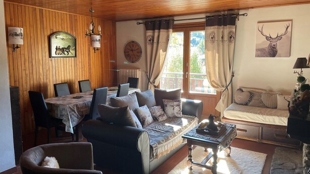 Een gezellige woonkamer met een houten plafond, een eethoek met een tafel en stoelen, een bank en een raam met gordijnen.