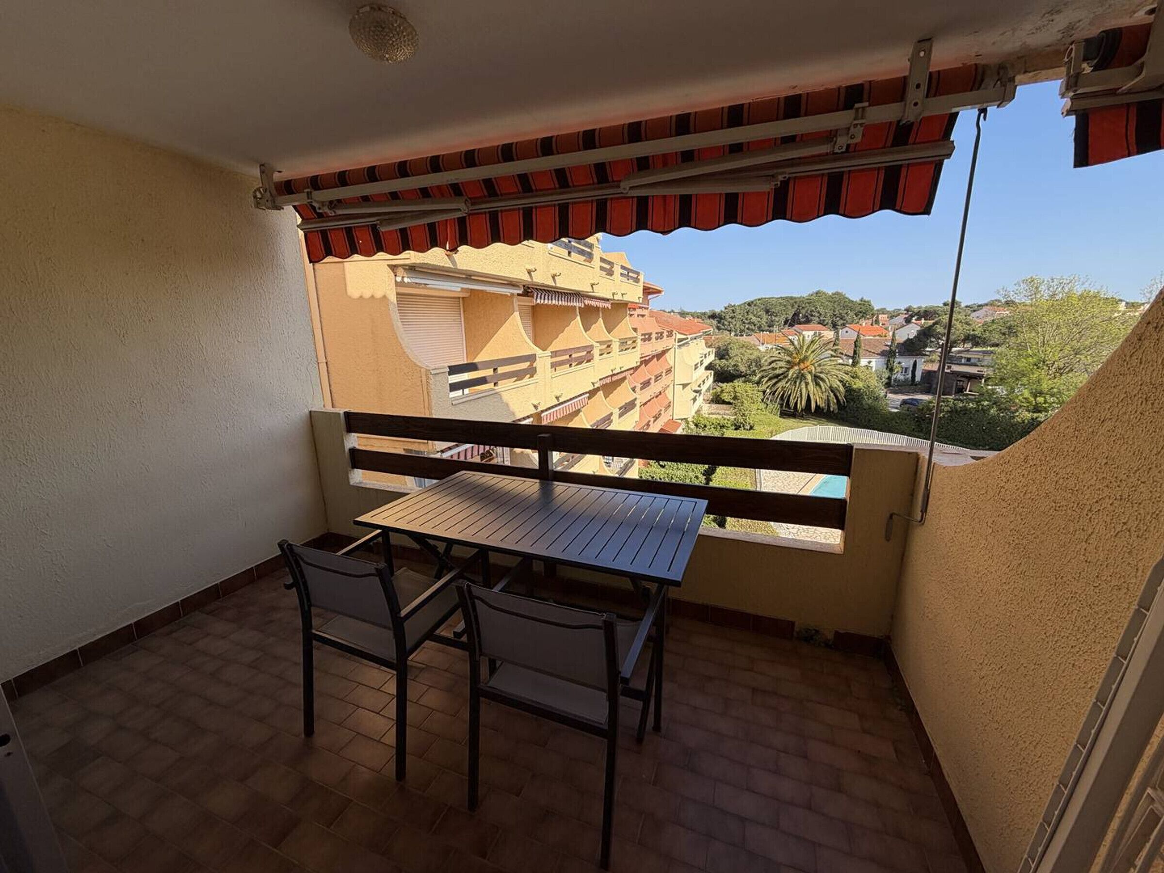 Un balcon avec une table et des chaises, un auvent rouge et une vue sur un quartier résidentiel avec des immeubles et de la verdure.