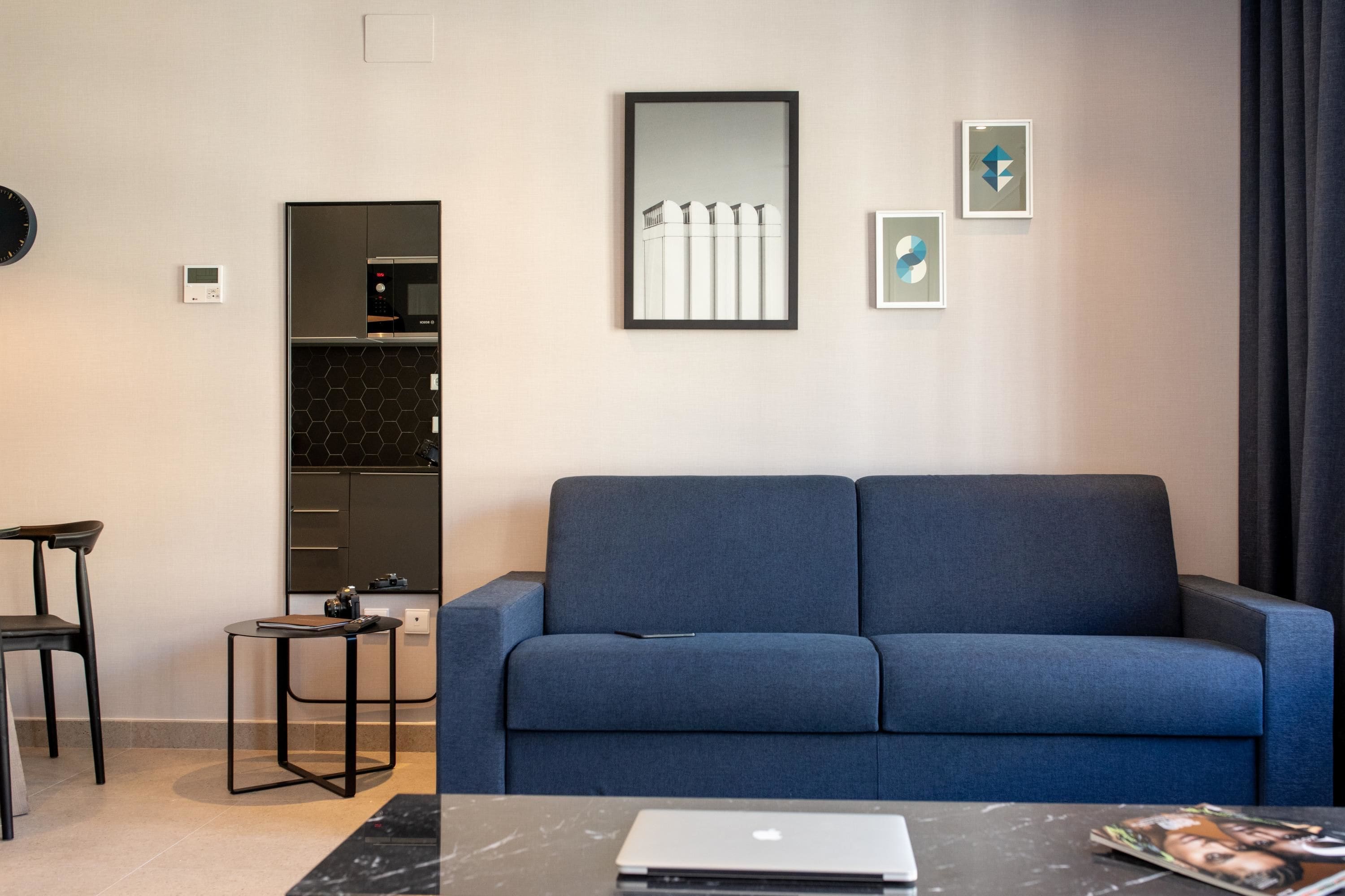 A modern living room with a blue sofa, a small table with a laptop, and a kitchen area with a microwave and cabinets.