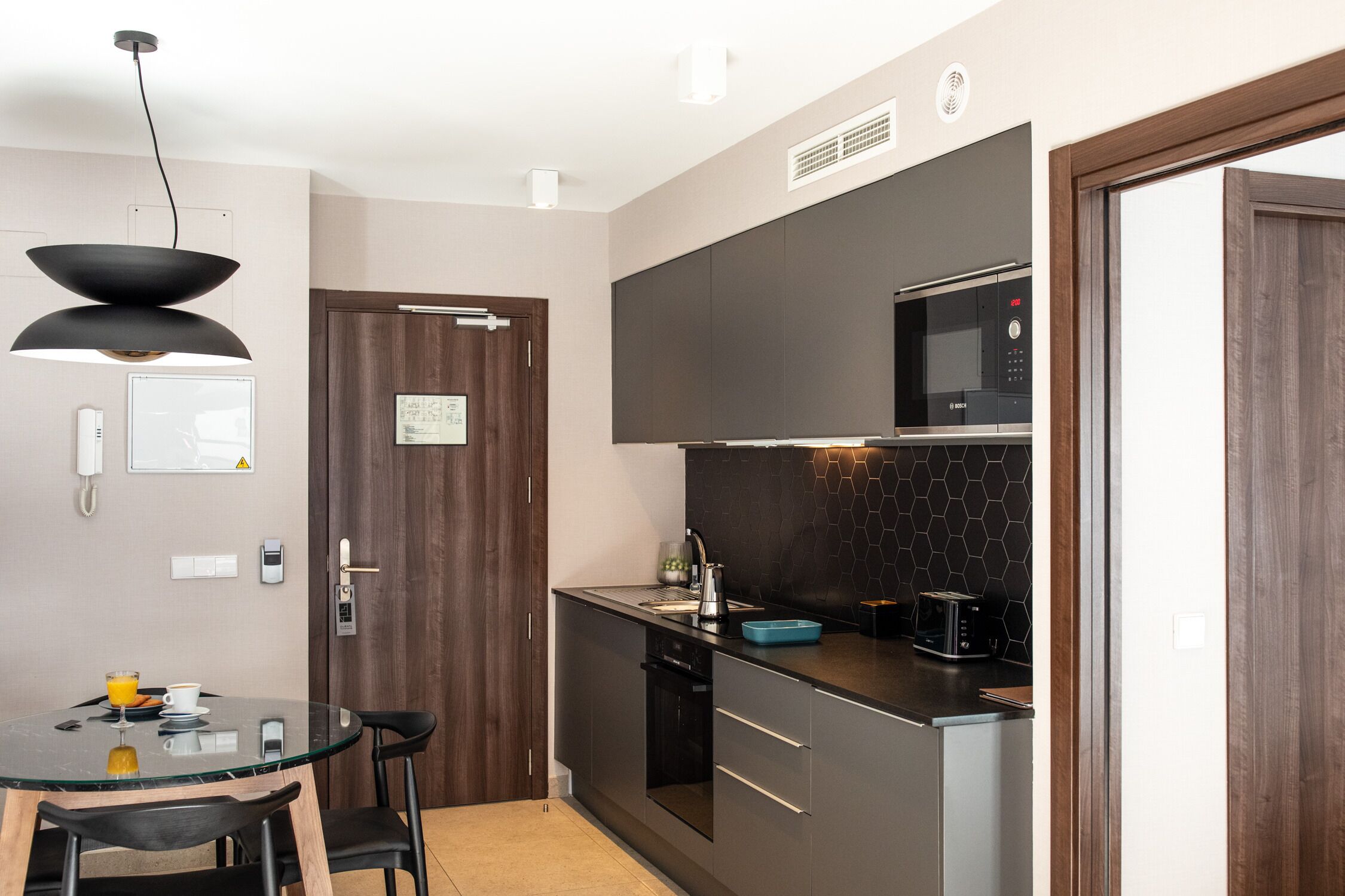 Una cucina compatta con mobili scuri, un forno a microonde, un tostapane e un tavolo in vetro con sedie.