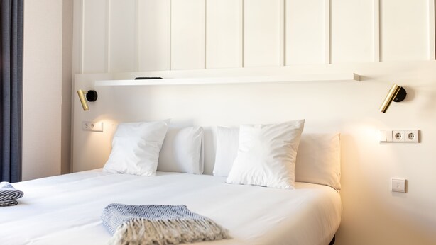 Un lit bien fait, avec du linge de lit blanc et un plaid, une table de chevet avec une lampe, et un luminaire fixé au mur.