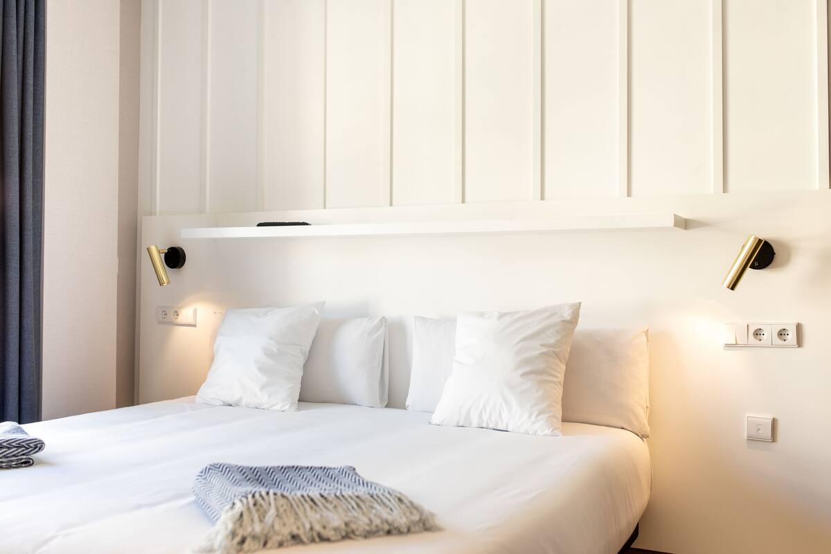 Una cama bien tendida con sábanas blancas y una manta decorativa, una mesita de noche con una lámpara, y un aplique en la pared.