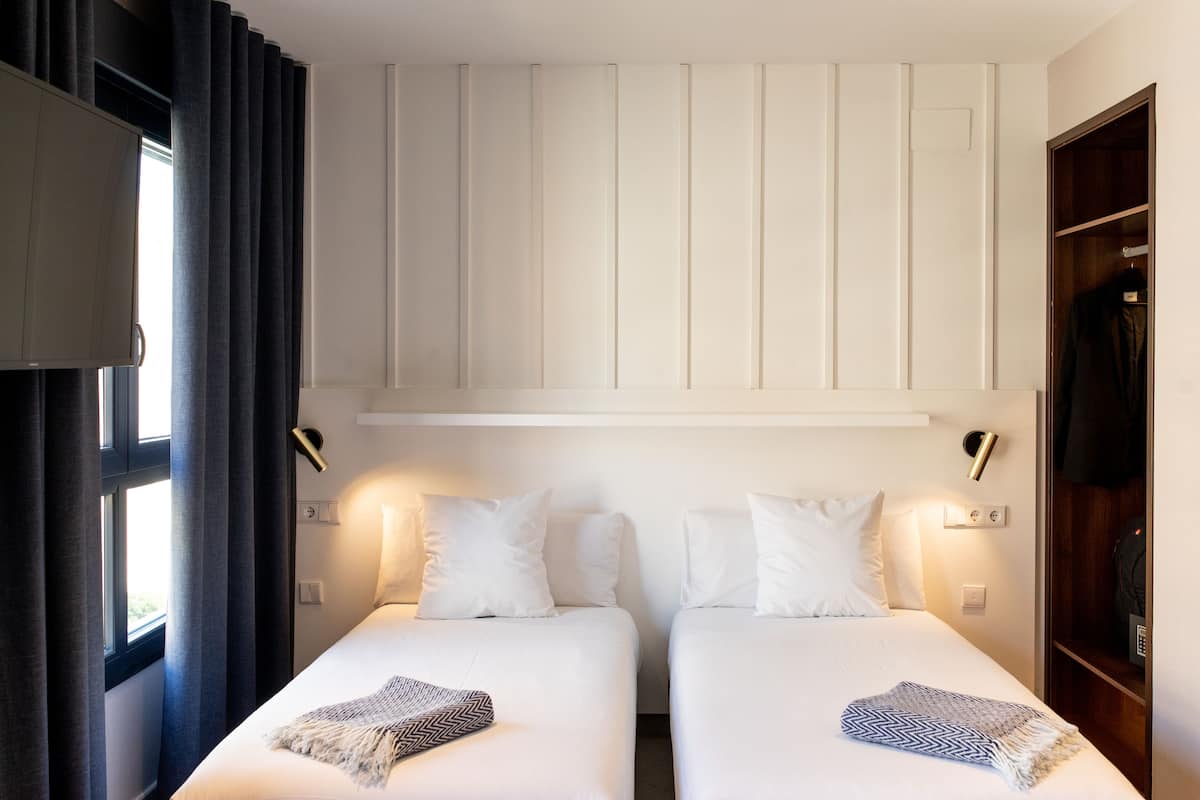 Una habitación de hotel con dos camas individuales, un armario, un espejo y una ventana con cortinas.