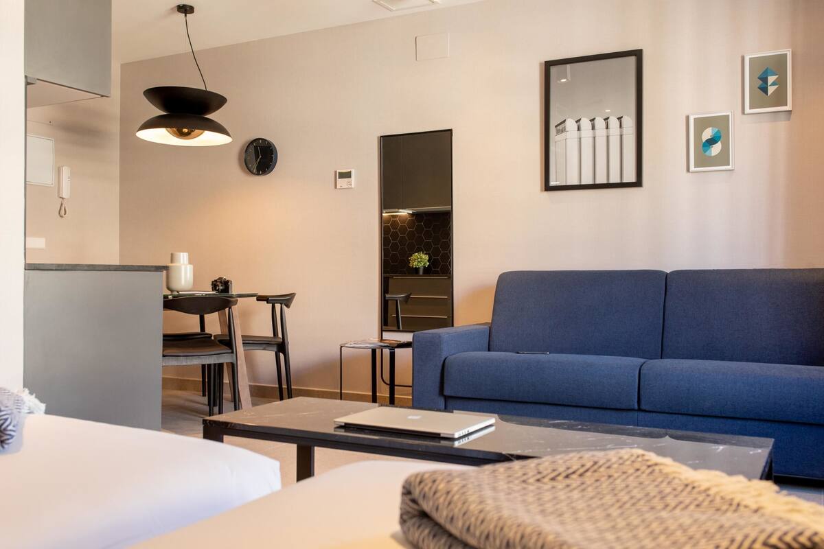 Una habitación de hotel moderna con un sofá azul, una mesa de comedor con sillas, una mesa auxiliar y un ordenador portátil sobre la mesa.