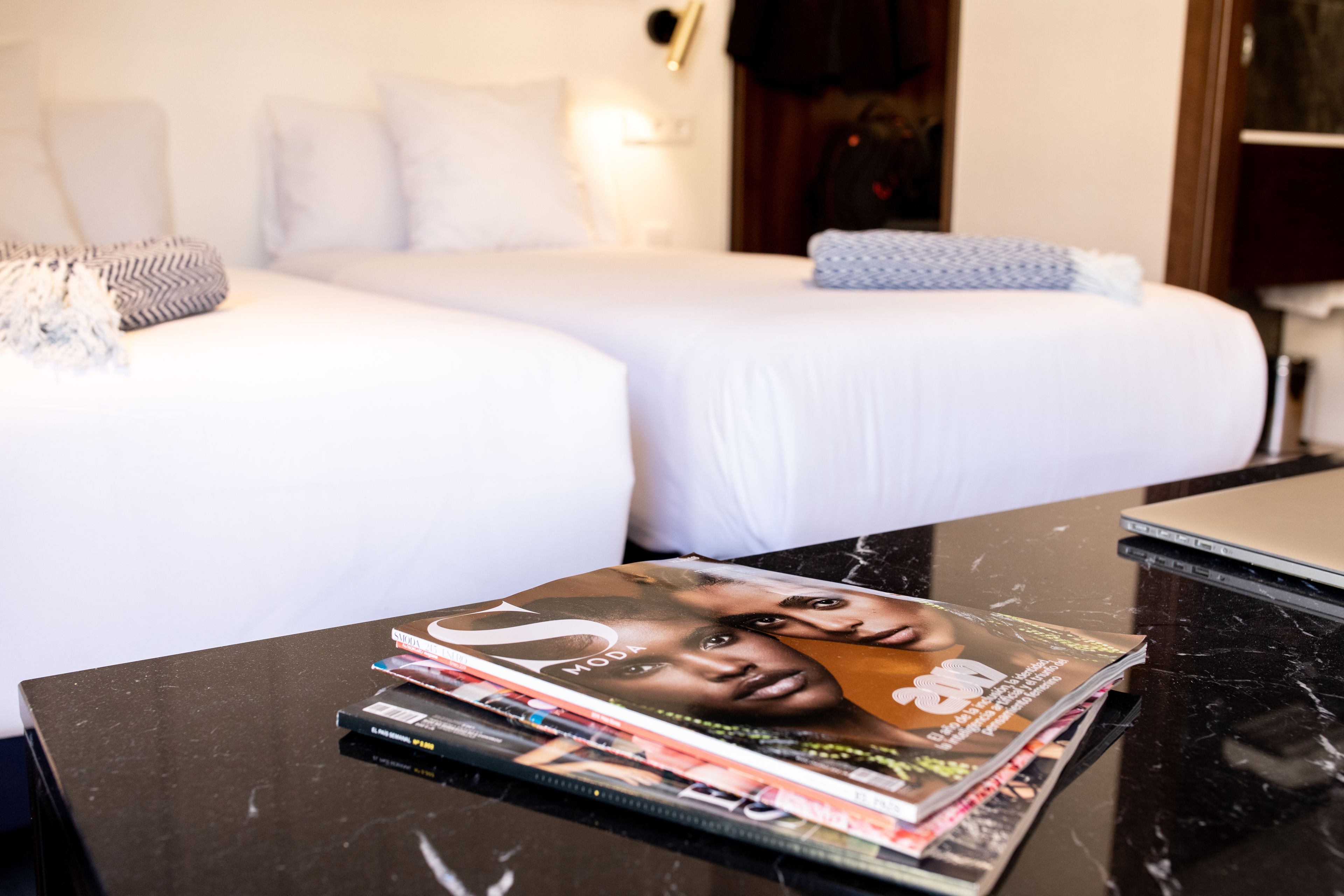 Habitació d'hotel amb un llit, una taula de nit amb revistes i un ordinador portàtil.