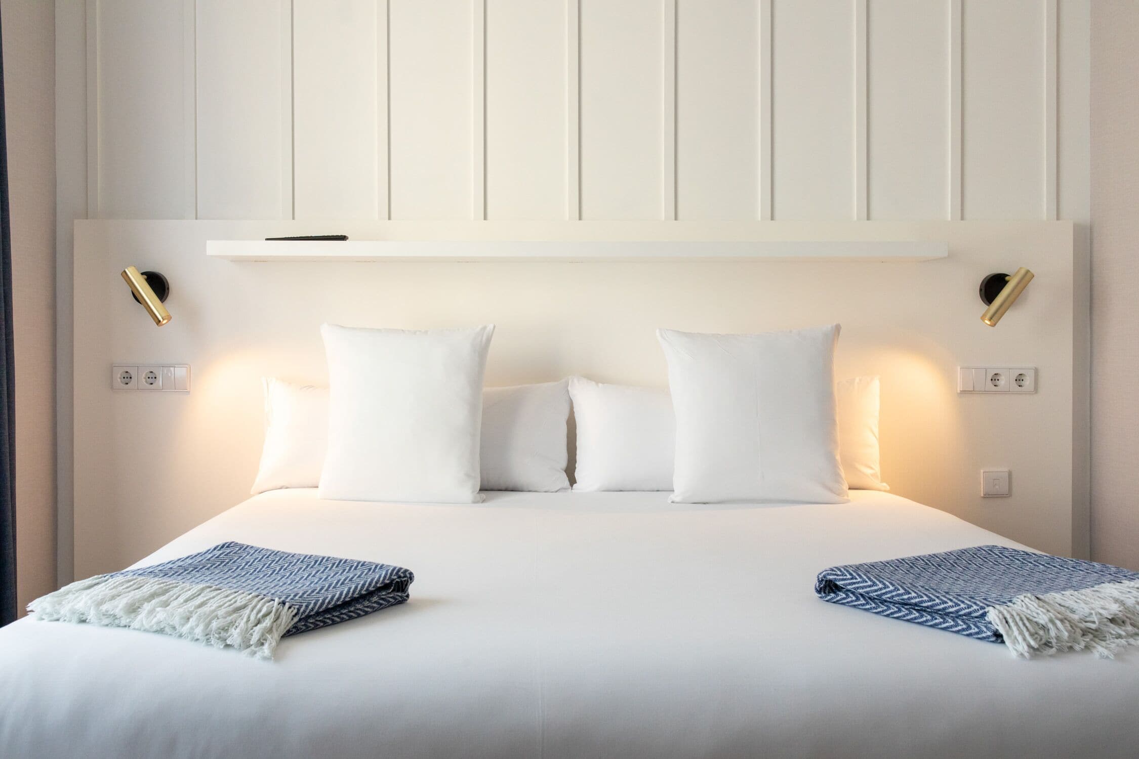 Una cama bien hecha con ropa de cama blanca y dos mantas decorativas, una mesita de noche con lámpara y un estante en la pared sobre la cama.
