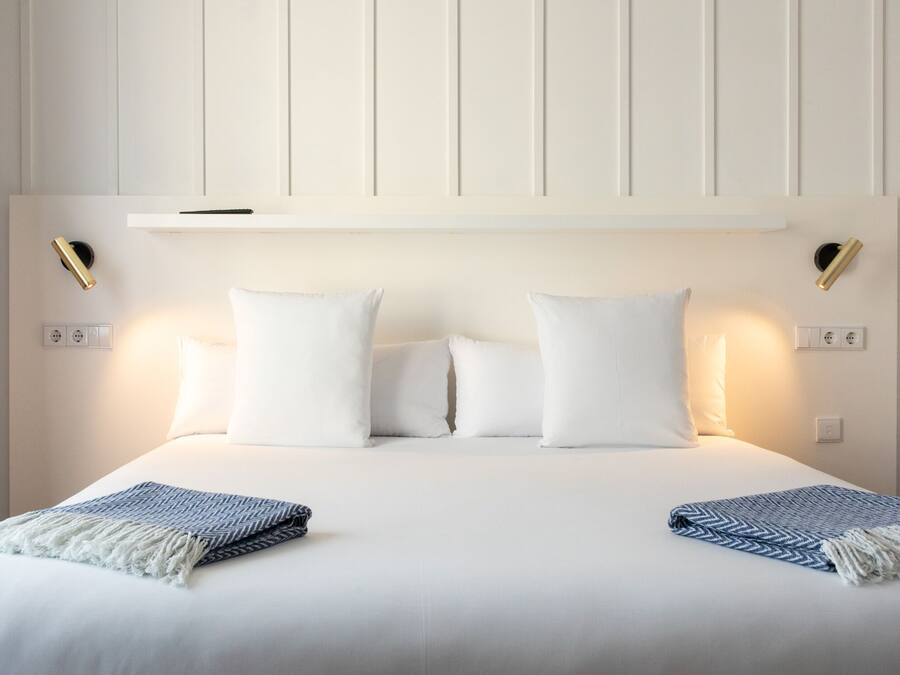 Una cama bien hecha con ropa de cama blanca y dos mantas decorativas, una mesita de noche con lámpara y un estante en la pared sobre la cama.