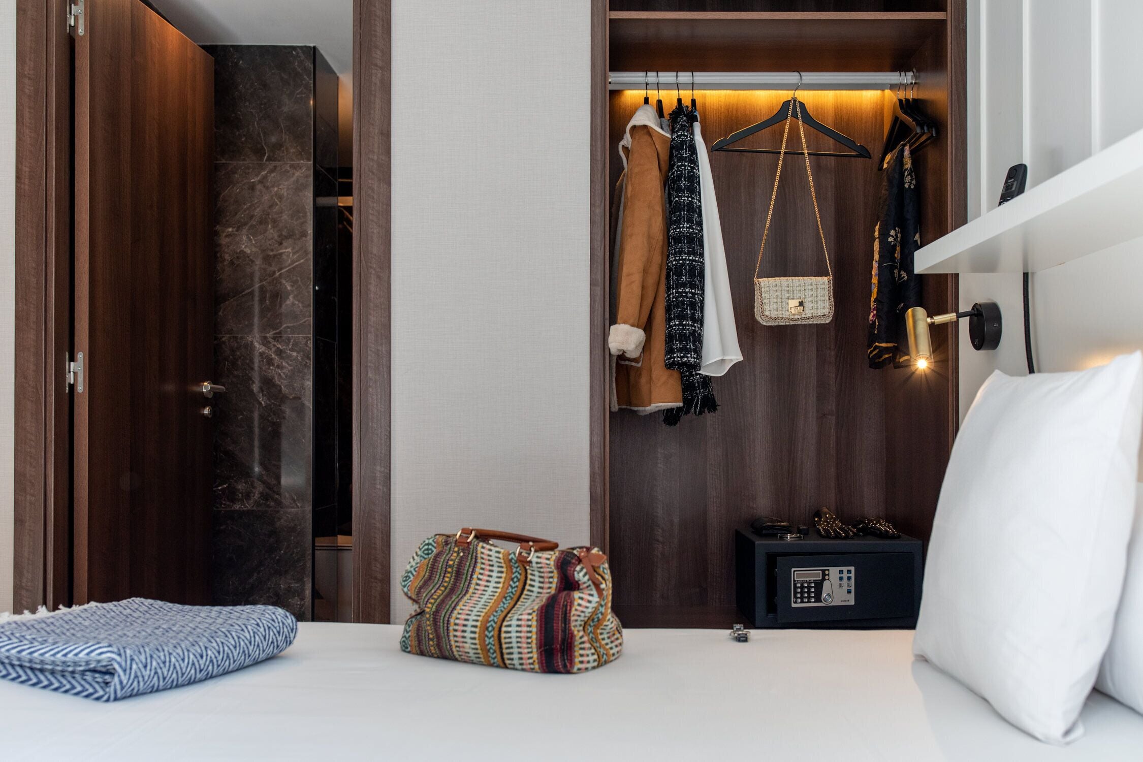 Quarto de hotel com cama, mesa de cabeceira, guarda-roupa e armário com roupas e bolsas.