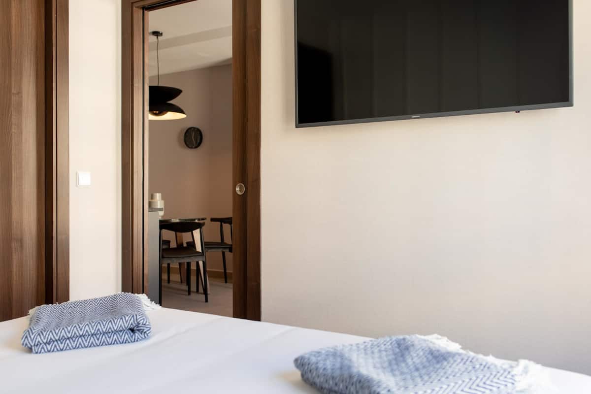 Habitación de hotel con cama, televisor en la pared, puerta a un comedor y un reloj a la vista.