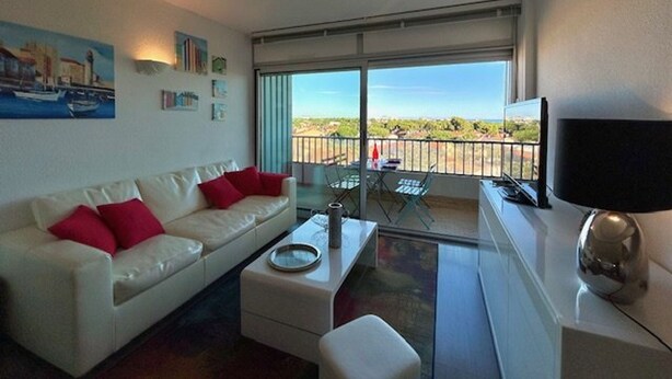 A modern living room with a white sofa, red cushions, a coffee table, and a TV on a stand. A balcony with a glass door leads to an outdoor area with a view of the landscape.