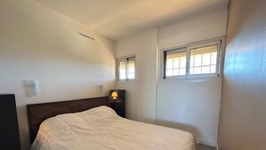 A bedroom with a single bed, a nightstand with a lamp, a window with blinds, and a wall-mounted light fixture.