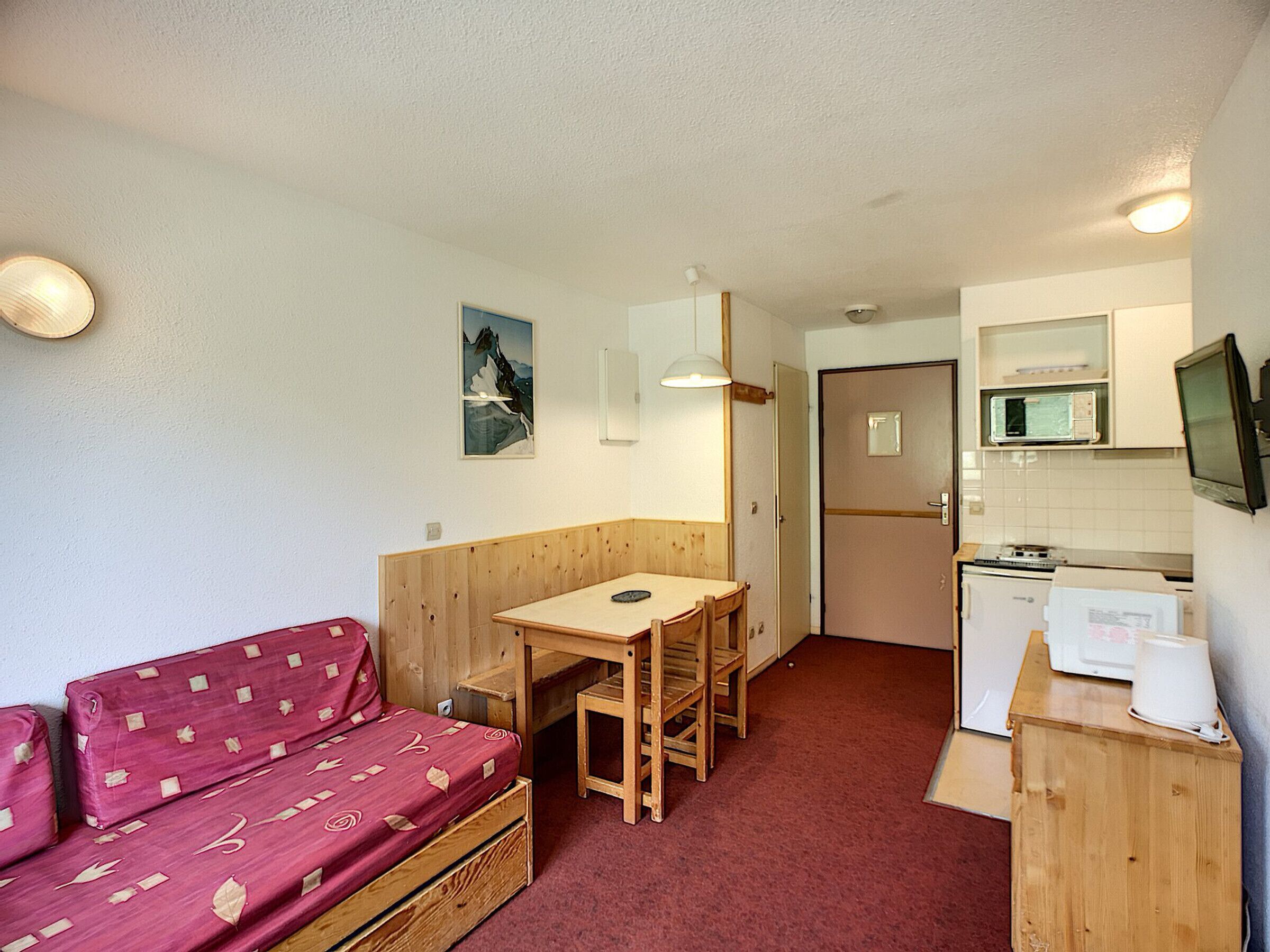 Een kleine kamer met een houten bankje, een tafeltje, een kitchenette en een rode, gestreept sofa.