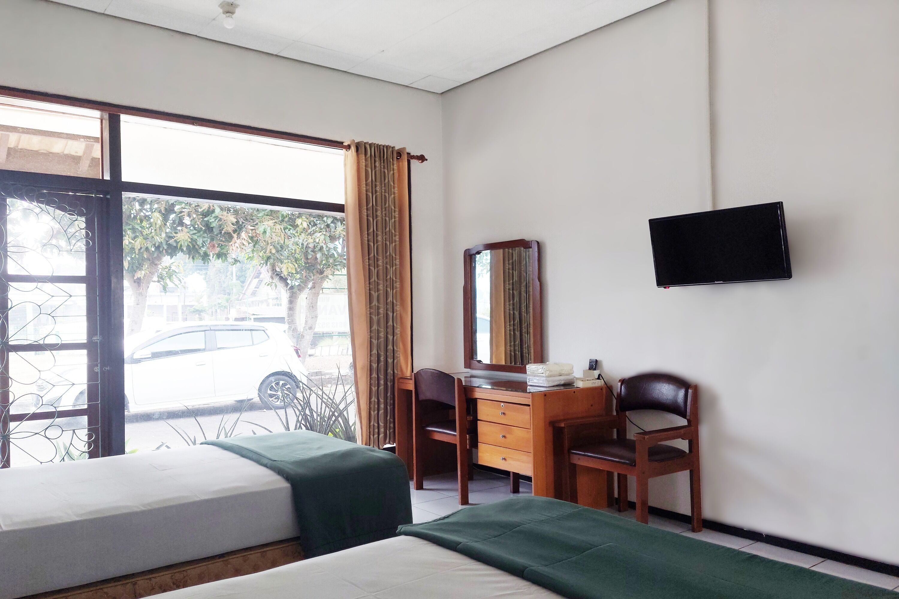 A hotel room with two beds, a wooden desk with a mirror, a television on the wall, and a window with curtains.