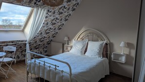 A bedroom with a bed, two bedside tables with lamps, a small table and chair, and a window with curtains. - Chambre Rose Blanche (Le Lude)