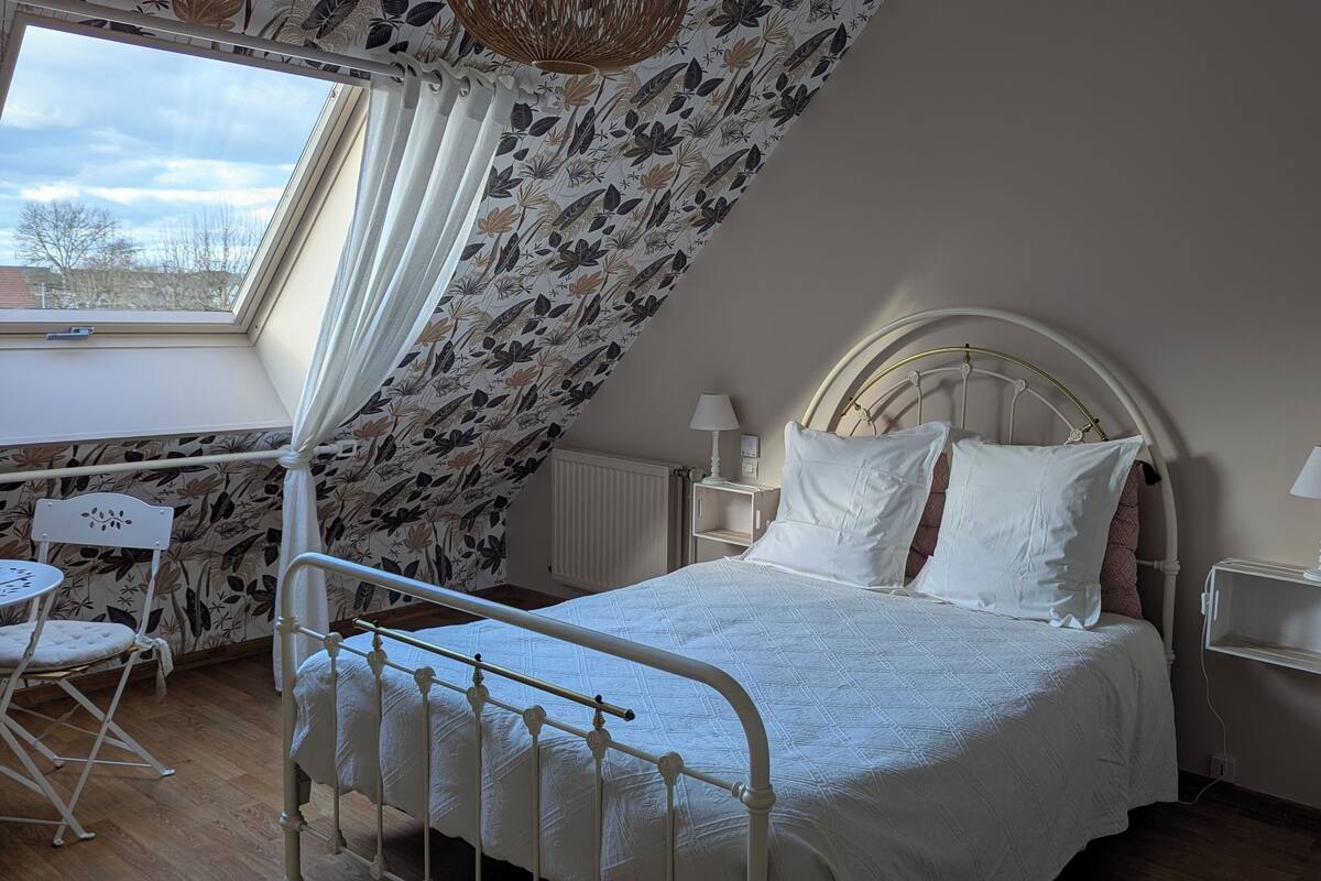 A bedroom with a bed, two bedside tables with lamps, a small table and chair, and a window with curtains.