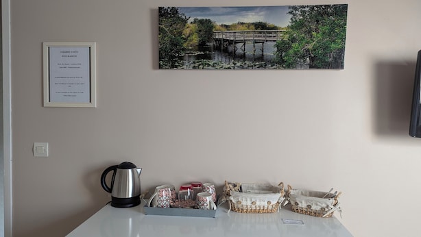 A hotel room with a framed welcome sign, a kettle, a tray with mugs and glasses, and a painting of a bridge over water. - Chambre Rose Blanche (Le Lude)