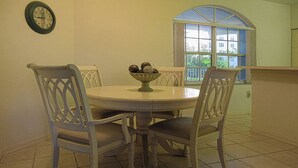 A dining area with a round table, four chairs, a bowl of fruit, a wall clock, and a window with a view of buildings.