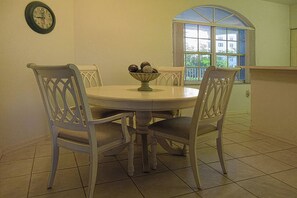 Un comedor con una mesa redonda, cuatro sillas, un frutero, un reloj de pared y una ventana con vistas a edificios.