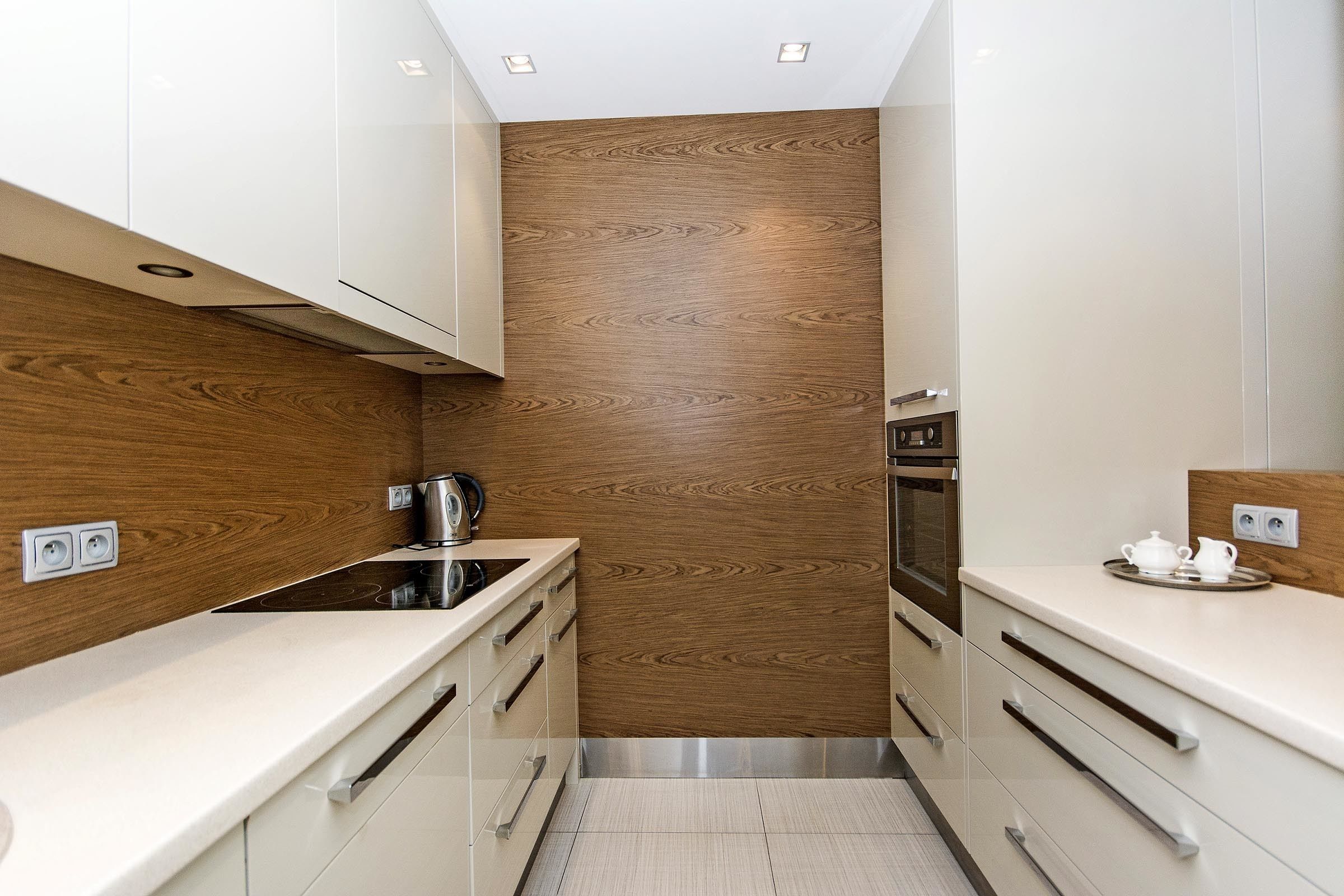 Cocina moderna con armarios blancos, un panel de madera decorativo, electrodomésticos de acero inoxidable y una pequeña zona de encimera con un hervidor y una tetera.