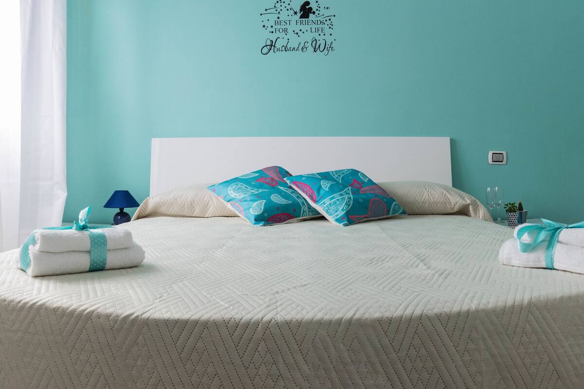 A neatly made bed with a patterned bedspread, two pillows with a paisley design, and two towels with a teal ribbon.