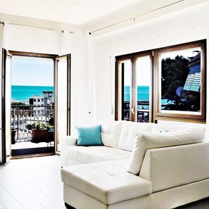 Un salon moderne avec un canapé blanc, un balcon offrant une vue sur la mer et une grande fenêtre donnant sur un immeuble et des arbres.