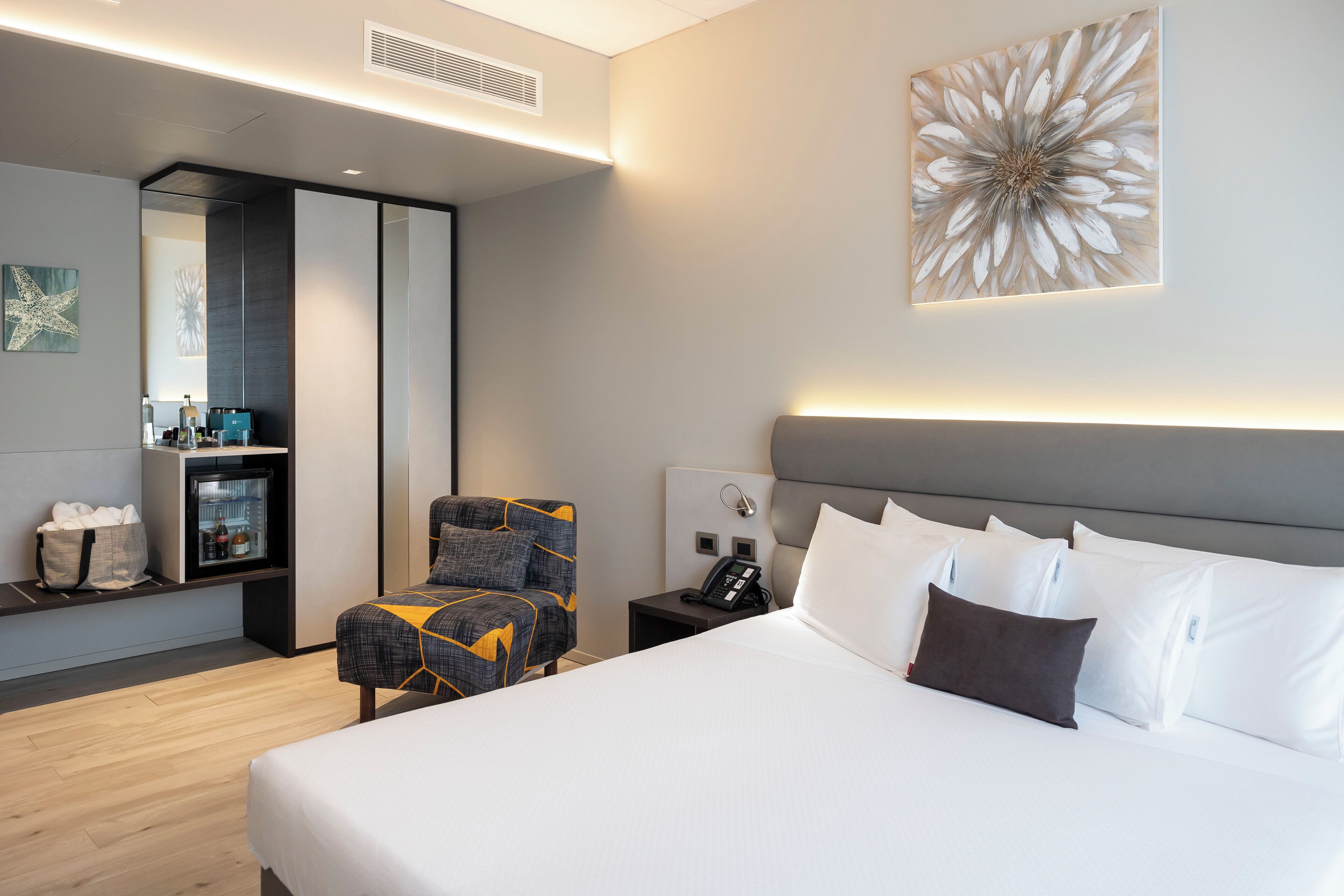 Una habitación de hotel moderna con una cama grande, una zona de estar con una silla, una mesita con un teléfono, un minibar y una obra de arte colgada en la pared.