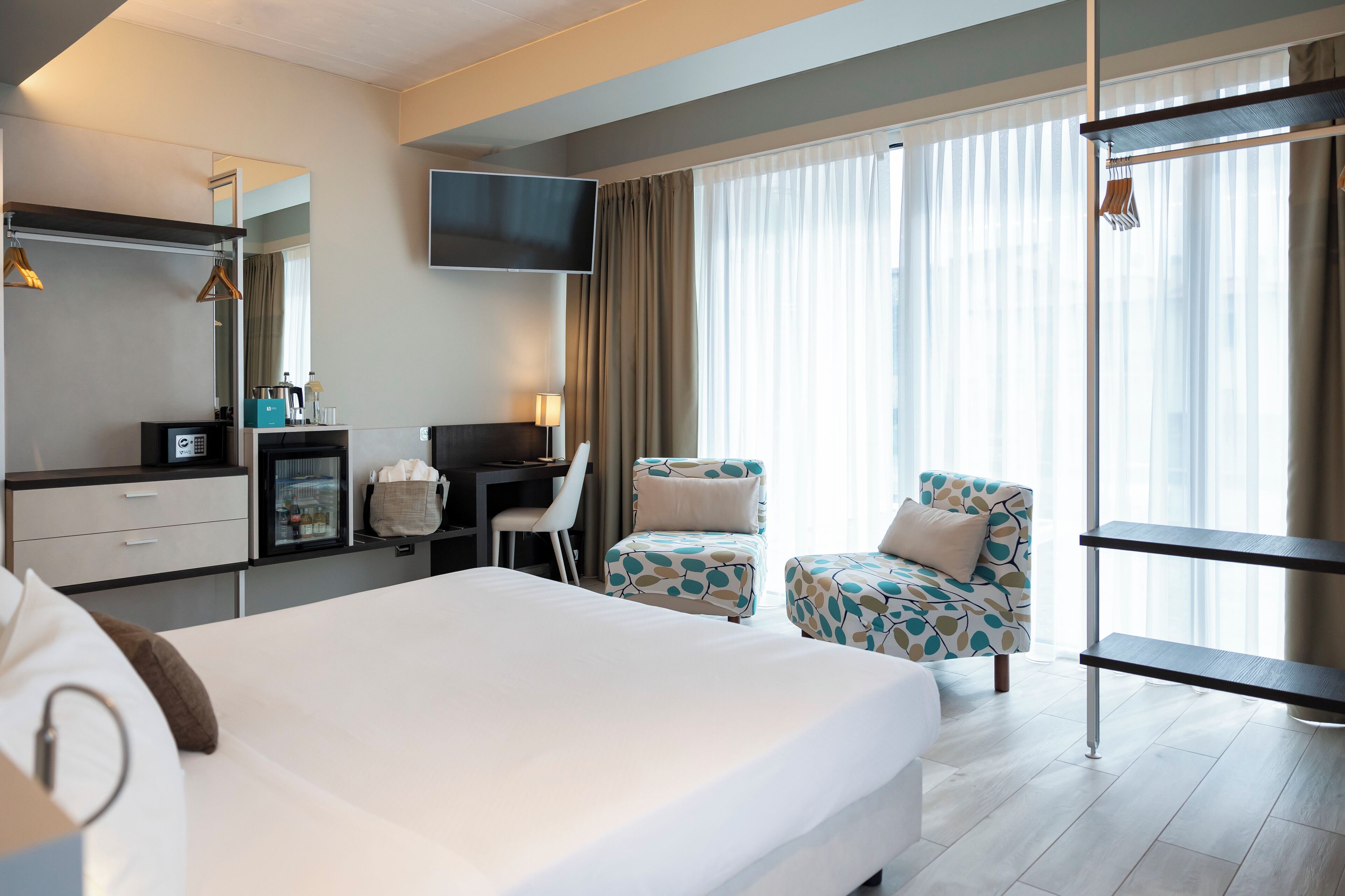 A hotel room with a large bed, a desk, a chair, a TV, and a window with curtains.