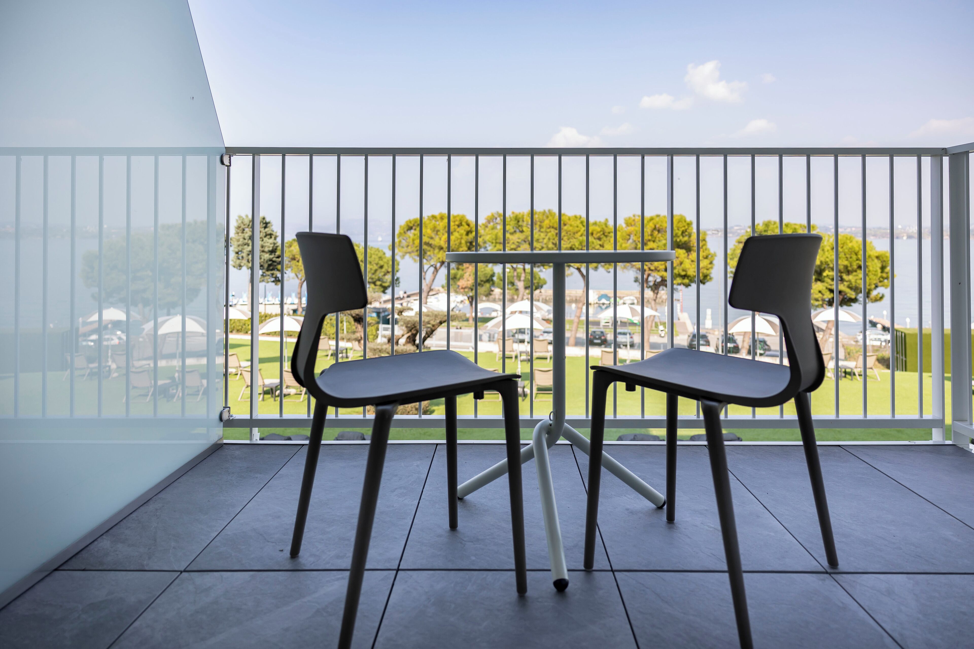 A balcony with two chairs and a table, overlooking a park and a body of water.