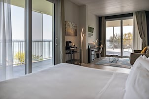 Habitación de hotel con una cama grande, un escritorio con silla, una mesita y un balcón con barandilla.