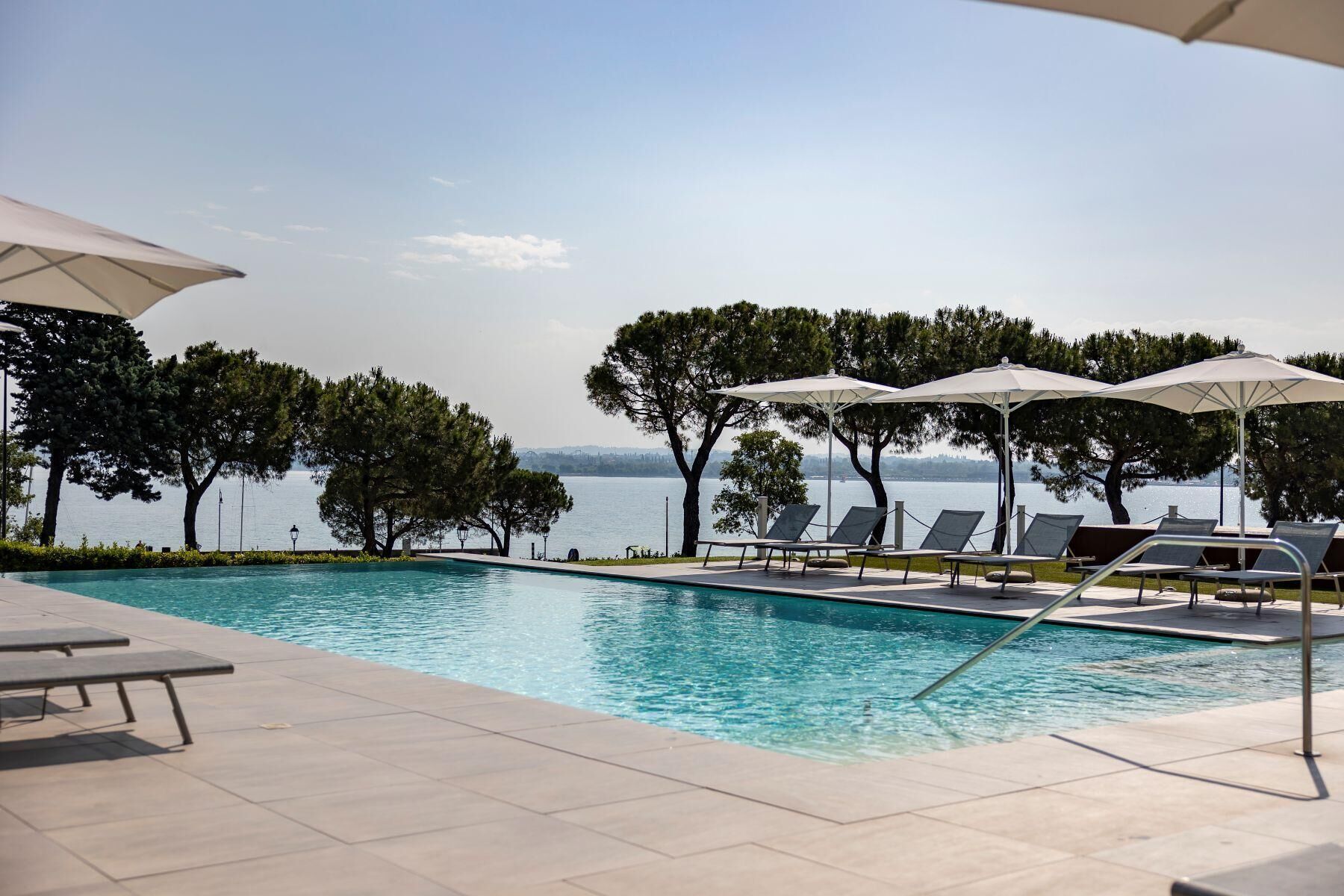 A pool area with lounge chairs and umbrellas, surrounded by trees and overlooking a body of water.