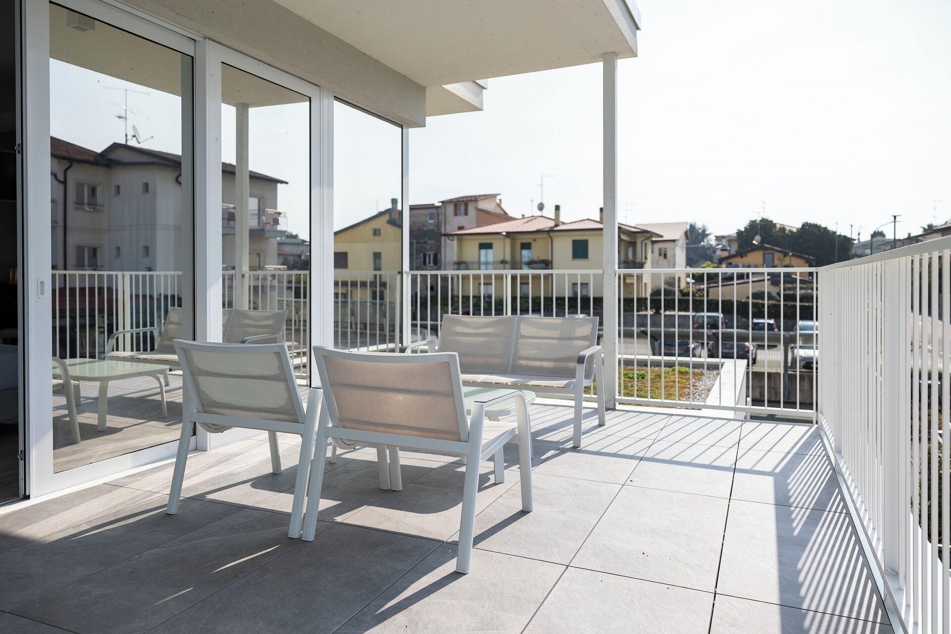 Un balcon avec des chaises blanches et une table, une porte vitrée et une vue sur des immeubles résidentiels.