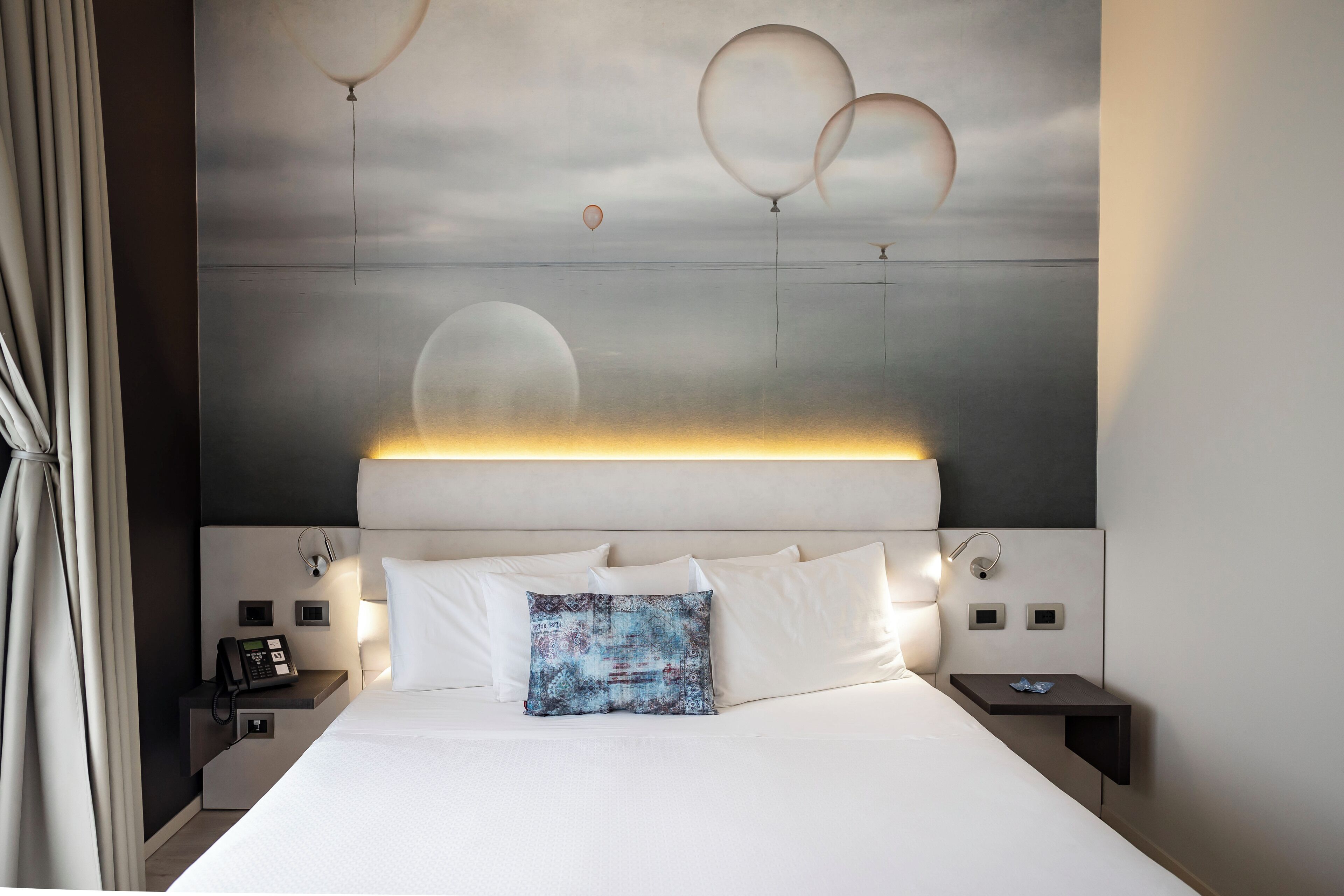 A modern hotel room with a large bed, a bedside table with a phone, and a wall-mounted light.