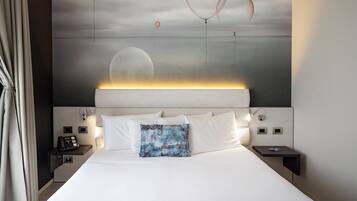 A modern hotel room with a large bed, a bedside table with a phone, and a wall-mounted light.