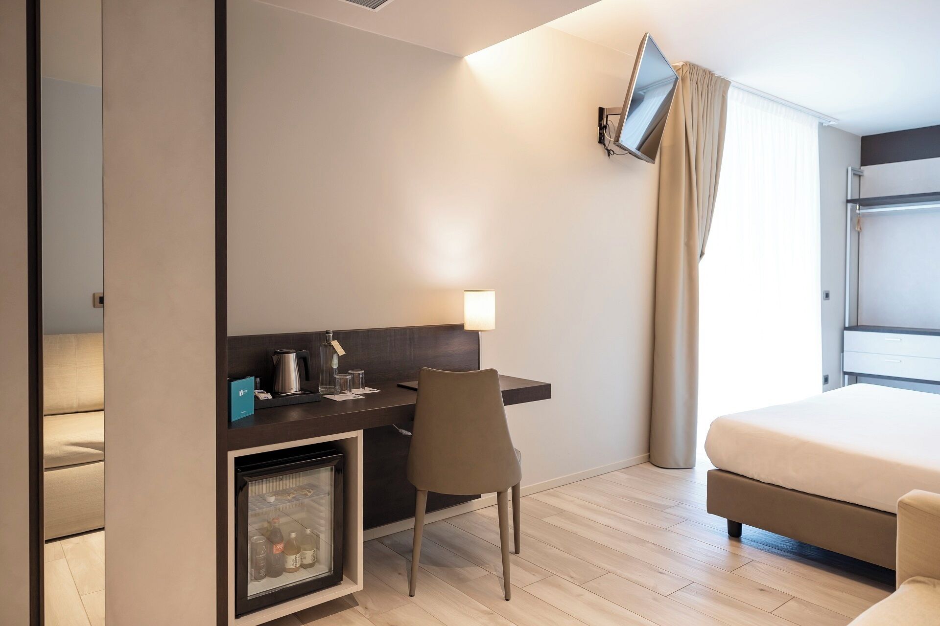 Ein Hotelzimmer mit Bett, Schreibtisch und Stuhl, kleinem Kühlschrank, Wand-Fernseher und einem Fenster mit Vorhängen.