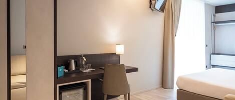 Habitación de hotel con cama, escritorio y silla, refrigerador pequeño, televisor de pantalla plana en la pared y ventana con cortinas.
