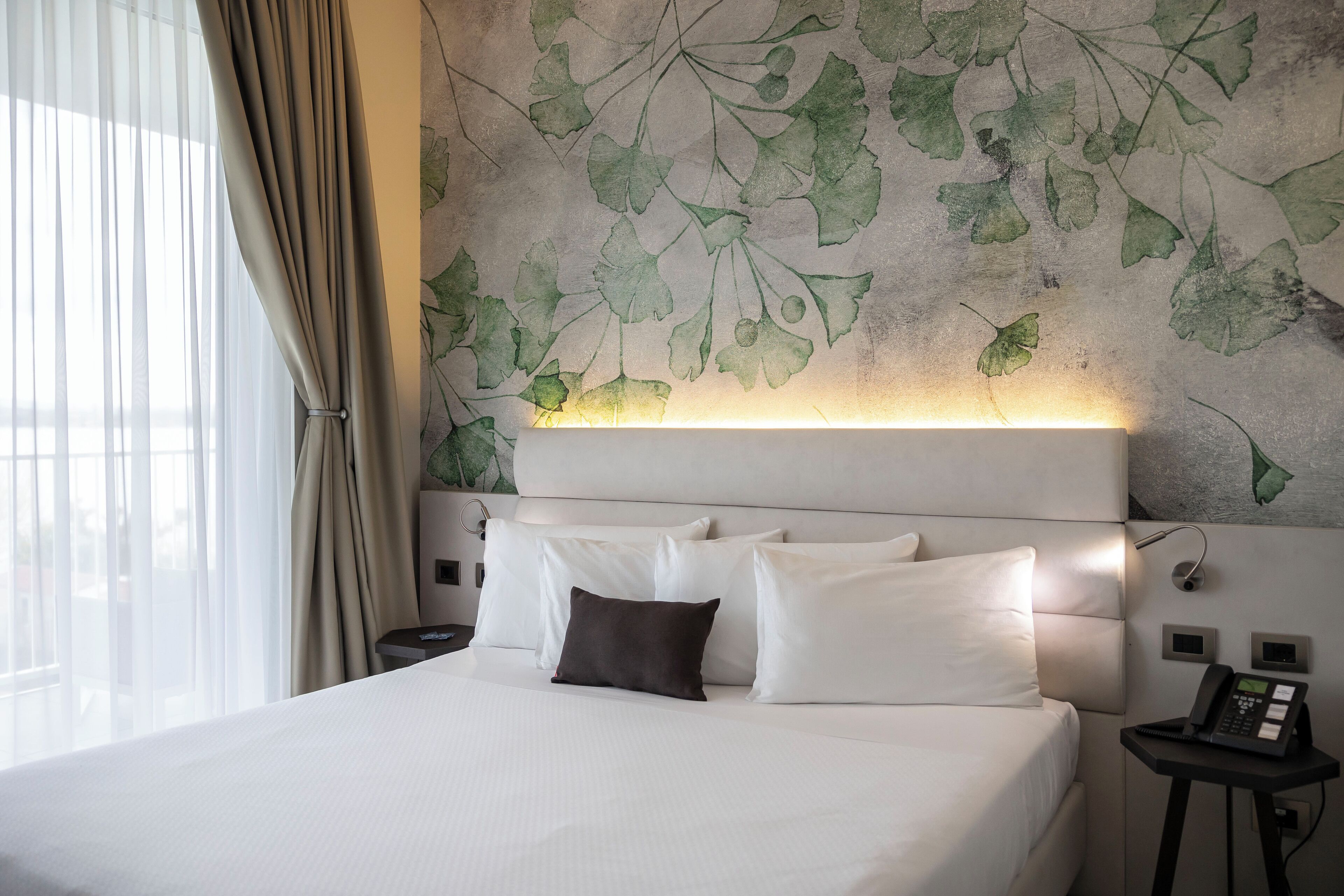 Habitación de hotel con cama, ropa de cama blanca, almohada marrón oscuro, teléfono en la pared y mesita de noche.
