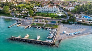 An aerial view of a coastal marina with boats docked, a pier, and a hotel with a pool in the background. - Vision Hotel (Peschiera del Garda)