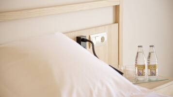 A neatly made bed with a wooden headboard, a bedside table with two bottles of water and a glass, and a power outlet on the wall.