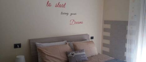 A bedroom with a bed, pillows, a bedside table, a lamp, and a wall with a motivational quote.